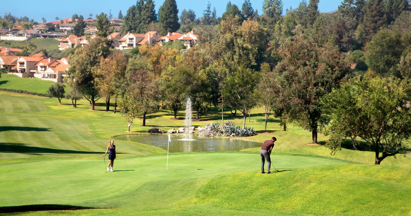 “Two people playing golf on a sunny course, with one putting on a green near a flagstick while another stands nearby, beside a small pond with a fountain and surrounded by trees and hillside homes.”